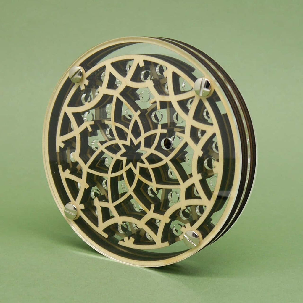 Seitlich aufgestelltes, rundes Kugellabyrinth aus hellem Holz und Acryl mit floral-sternförmigem Muster; vier Metallschrauben halten die transparente Front, silberne Kugeln schimmern im Inneren vor grünem Hintergrund.