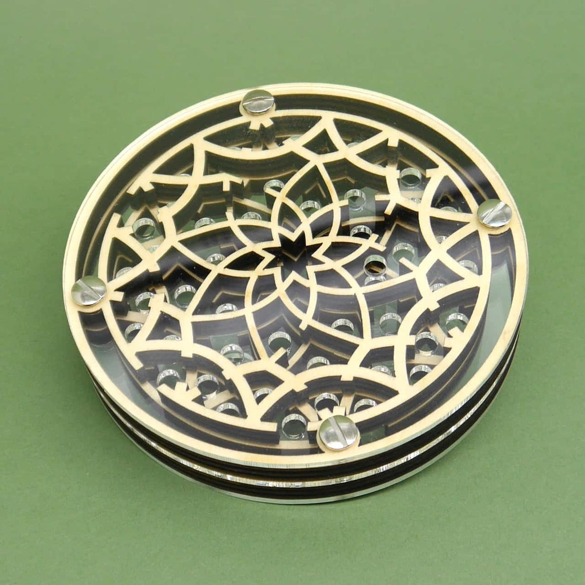 Schräger Blick von oben auf das flach liegende, runde Geduldsspiel: ein feingefrästes Blütenlabyrinth aus Holz unter einer klaren Deckscheibe, fixiert von vier Schrauben; kleine Metallkugeln blitzen zwischen den Bahnen, grüner Untergrund.