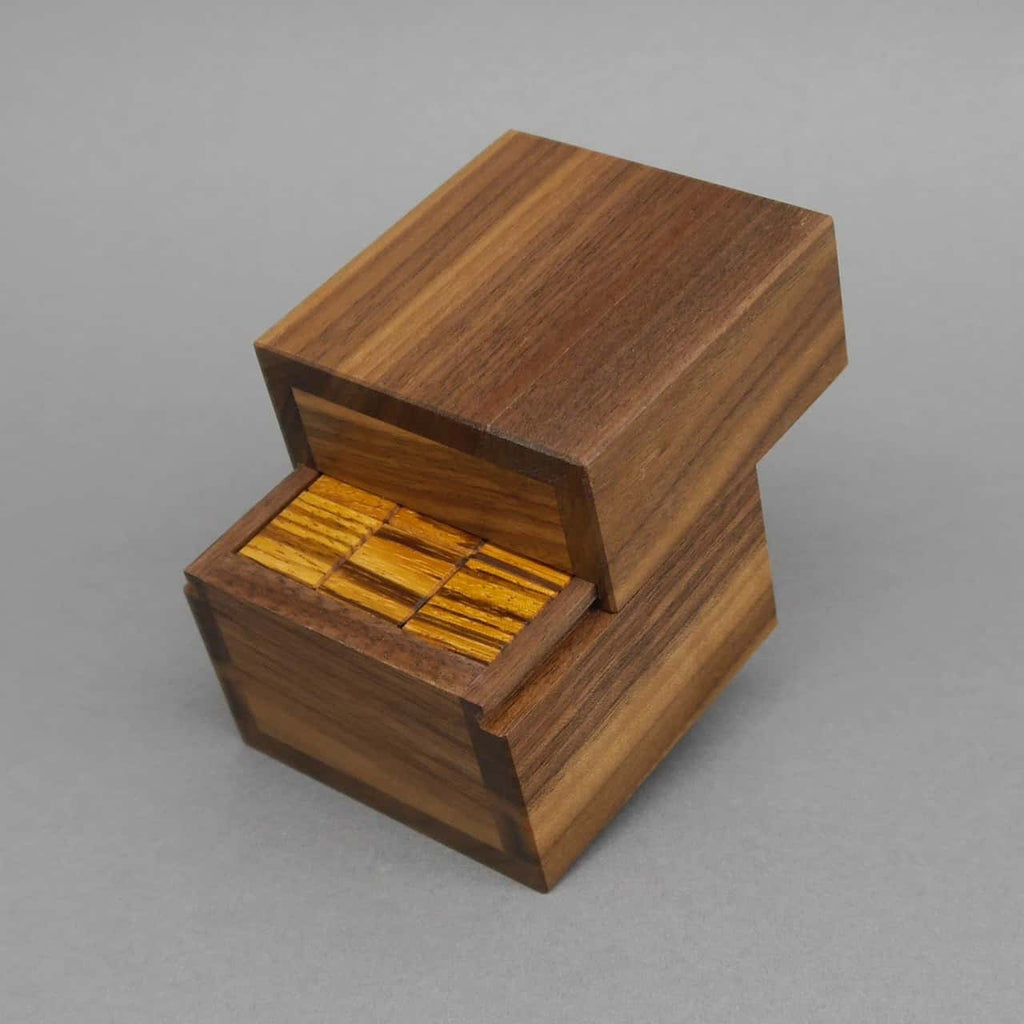 Geöffnetes Denkspiel aus dunkel gemasertem Nussbaumholz; der Deckel ist hochgeklappt und zeigt gelb gestreifte Puzzleteile im Inneren.