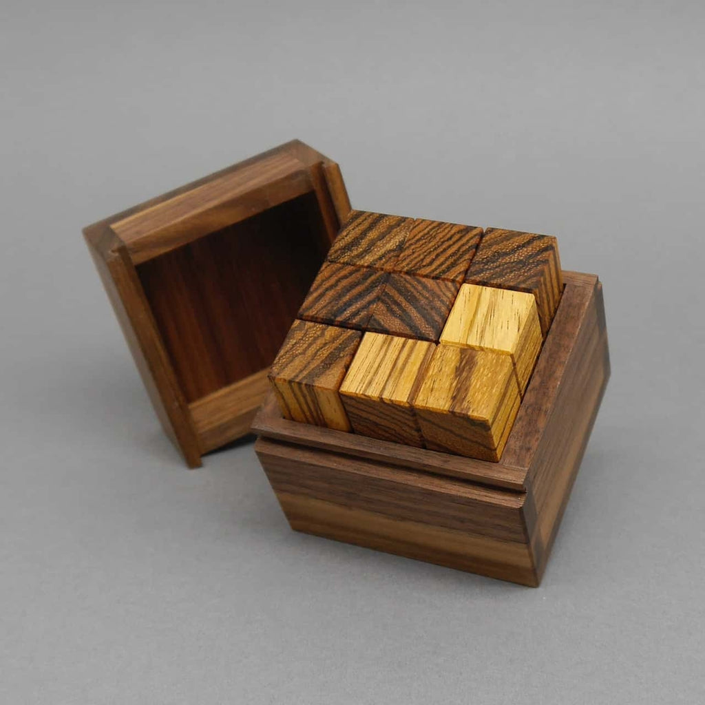 Fast fertig gesetztes IQ-Puzzle: kontrastreich gemaserte Würfel füllen eine offene Holzbox, der Deckel lehnt seitlich an.