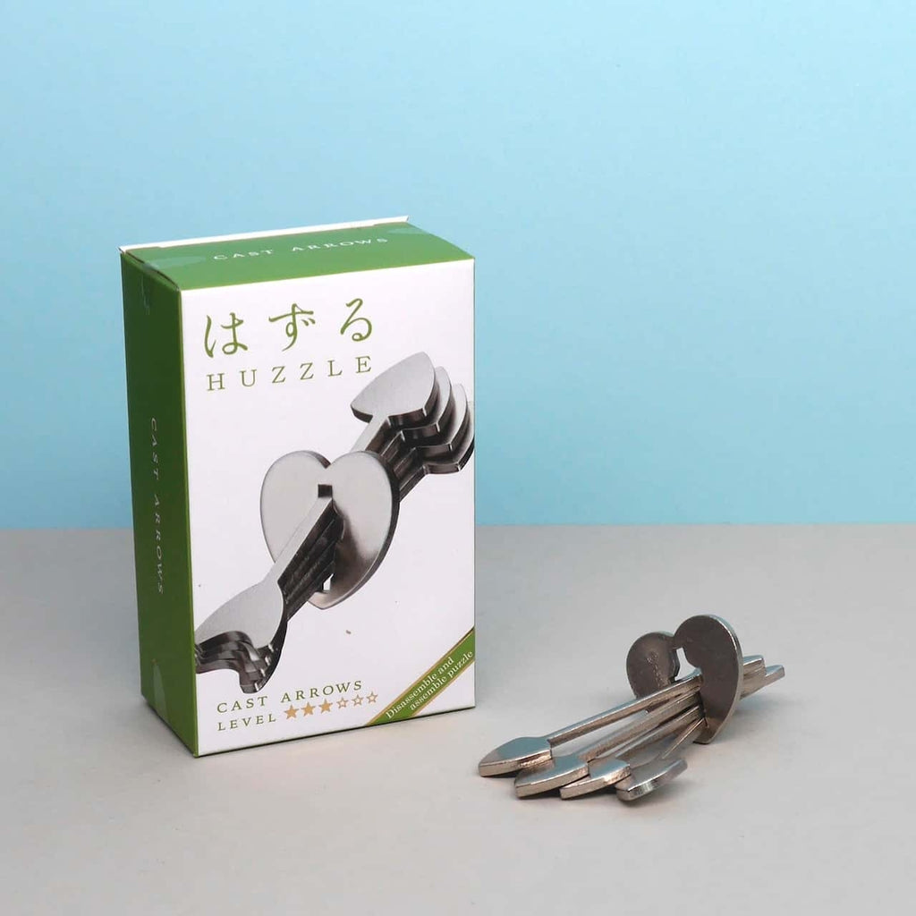 Huzzle Puzzle: Verpackung und Metallpuzzle mit pfeilförmigen Teilen auf neutralem Hintergrund.