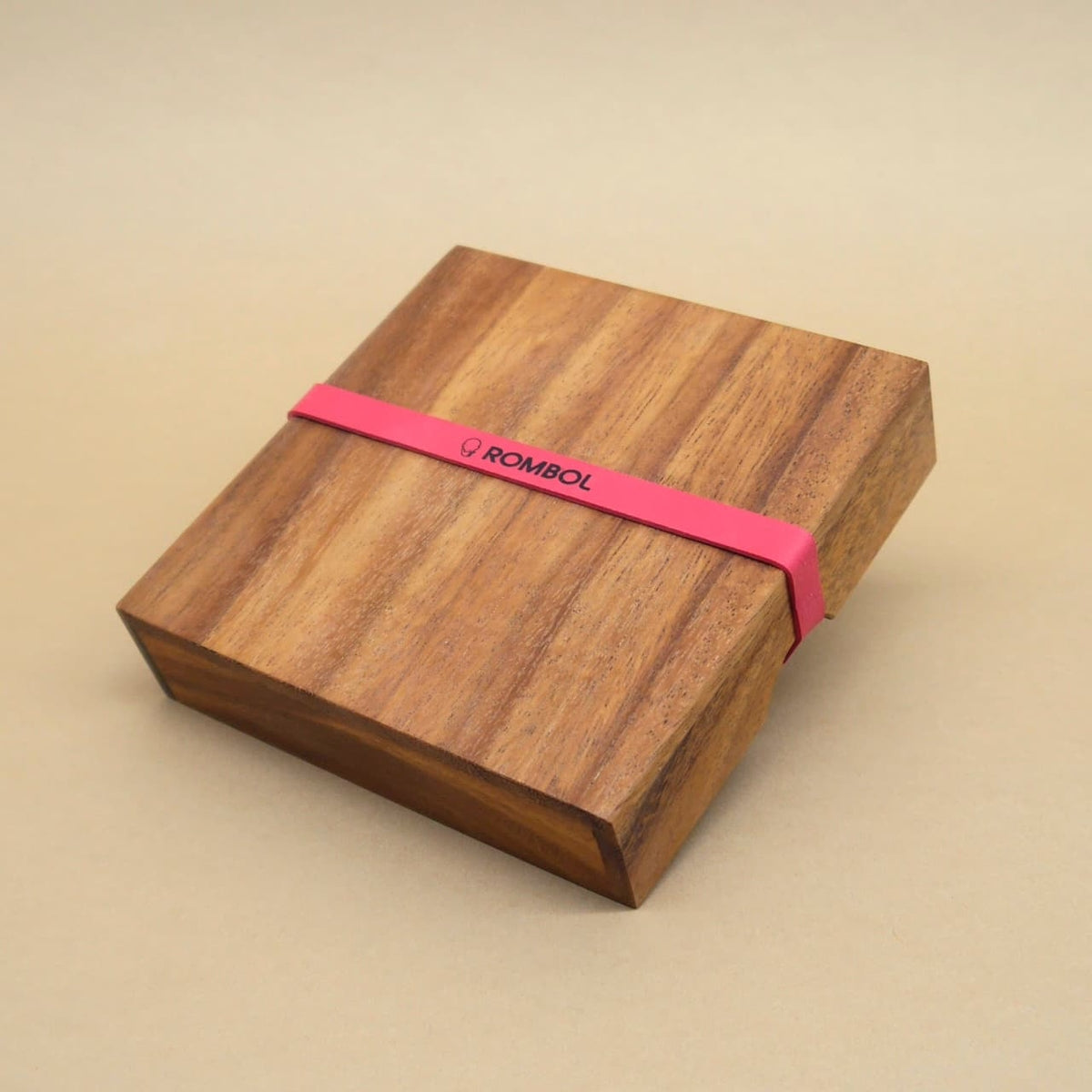 Geschlossene, quadratische Holzbox eines Knobelspiels, von einem rotem Band mit ROMBOL-Logo umspannt.