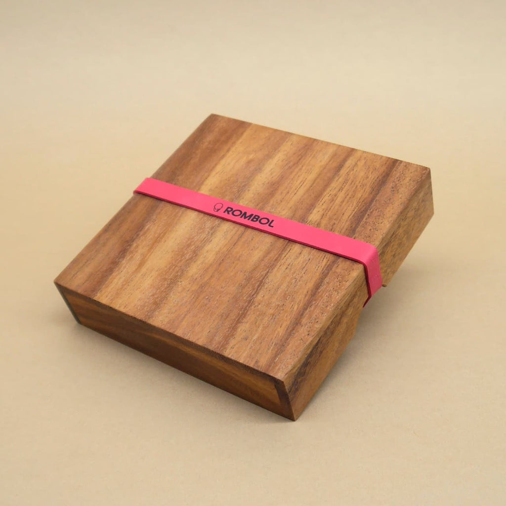Geschlossene, quadratische Holzbox eines Knobelspiels, von einem rotem Band mit ROMBOL-Logo umspannt.