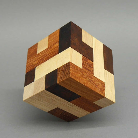 Zusammengebauter Holzwürfel aus hell-, mittel- und dunkelbraunen Leisten balanciert diagonal auf einer Ecke vor grauem Hintergrund.