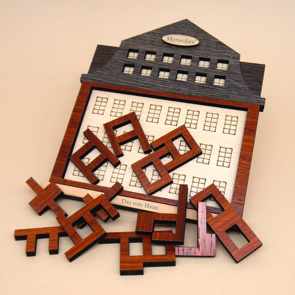 Einzelteile des Holzpuzzles ‚Das rote Haus‘ liegen verstreut auf einer Oberfläche; das rote Hauselement ist deutlich erkennbar.