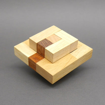 Das Holz‑Denkspiel ist fertig zu einem zweistufigen, quadratischen Gebilde aus hellen und dunklen Quadern zusammengesetzt und schwebt leicht über dem Untergrund.