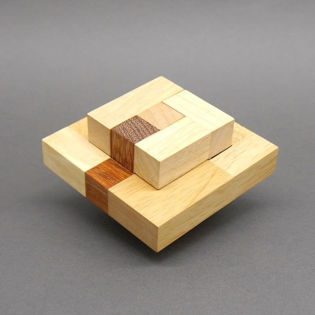 Das Holz‑Denkspiel ist fertig zu einem zweistufigen, quadratischen Gebilde aus hellen und dunklen Quadern zusammengesetzt und schwebt leicht über dem Untergrund.
