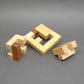 Drei zerlegte Elemente eines Holz‑Denksport‑Puzzles – ein quadratischer Rahmen und zwei blockartige Teile – liegen vor einer grauen Fläche.