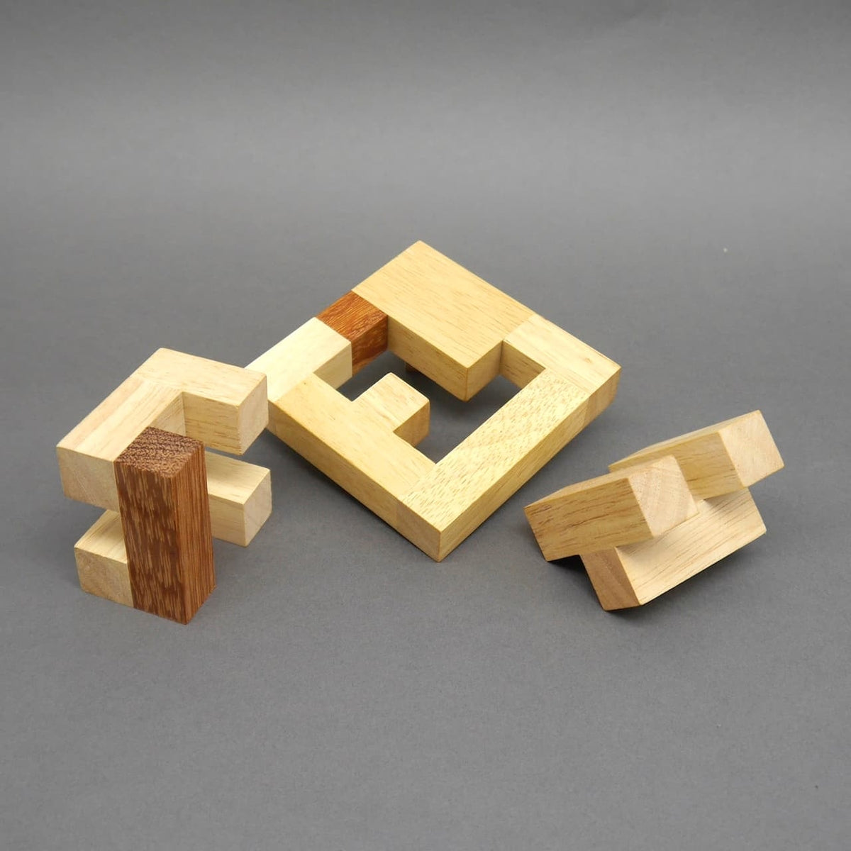 Drei zerlegte Elemente eines Holz‑Denksport‑Puzzles – ein quadratischer Rahmen und zwei blockartige Teile – liegen vor einer grauen Fläche.
