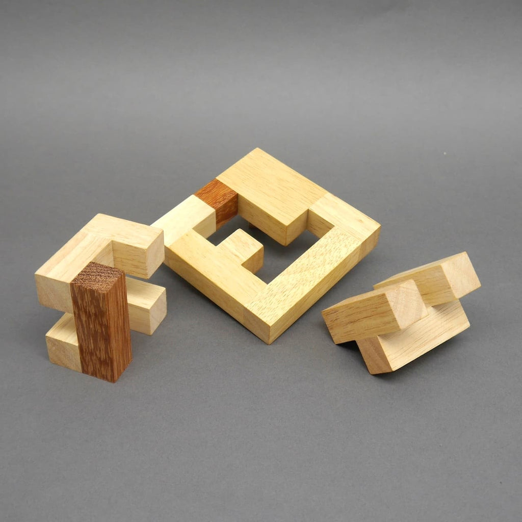 Drei zerlegte Elemente eines Holz‑Denksport‑Puzzles – ein quadratischer Rahmen und zwei blockartige Teile – liegen vor einer grauen Fläche.