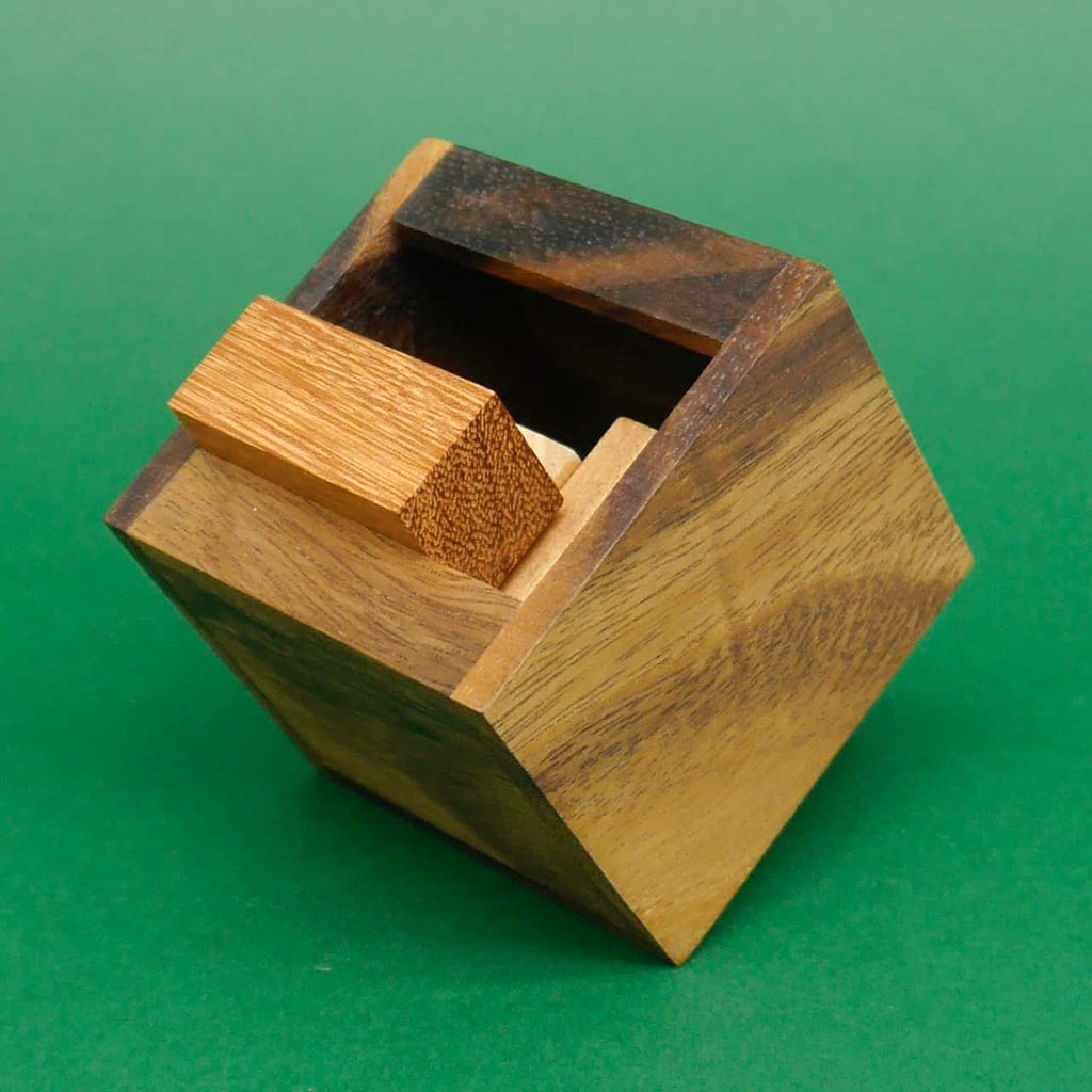 Holz-Brainteaser: Ein schräg stehendes Würfelkästchen zeigt einen halb eingesteckten rechteckigen Holzstein vor grünem Hintergrund.