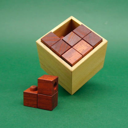 Eine fast vollständig gefüllte Holzkiste zeigt sauber angeordnete Würfelblöcke; das letzte Teil des Knobelpuzzle liegt griffbereit davor.