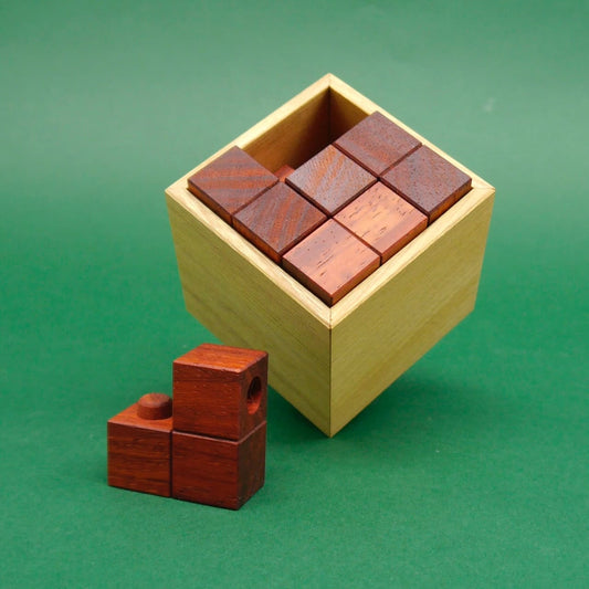 Eine fast vollständig gefüllte Holzkiste zeigt sauber angeordnete Würfelblöcke; das letzte Teil des Knobelpuzzle liegt griffbereit davor.