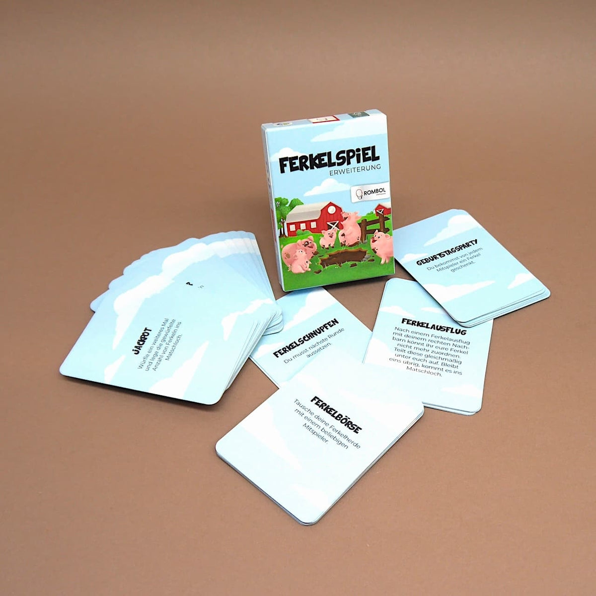 Mehrere hellblaue Spielkarten der „Ferkelspiel“-Erweiterung sind fächerförmig um die stehende Box auf einem braunen Untergrund ausgelegt.