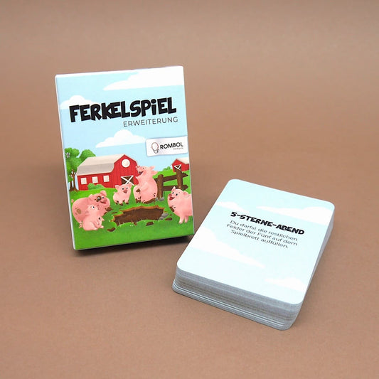 Die aufrecht stehende „Ferkelspiel“-Erweiterungsbox zeigt fröhliche Schweinchen vor einem Bauernhof; rechts daneben liegt ein ordentlicher Stapel Karten.