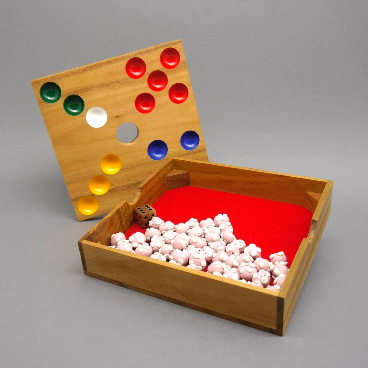 Geöffnetes Brettspiel „Ferkelspiel“: herausgenommene Spielplatte mit farbigen Mulden lehnt an der Holzkiste, innen rotes Filzfach voller Schweinchen und ein Holzwürfel.
