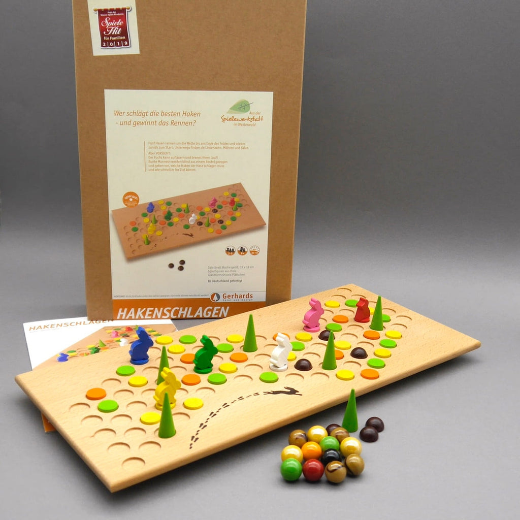 Aufgebautes Taktikspiel Hakenschlagen: Holzbrett mit bunten Scheiben, Kegeln wie Bäumchen, Hasenfiguren und Murmeln vor der Box.