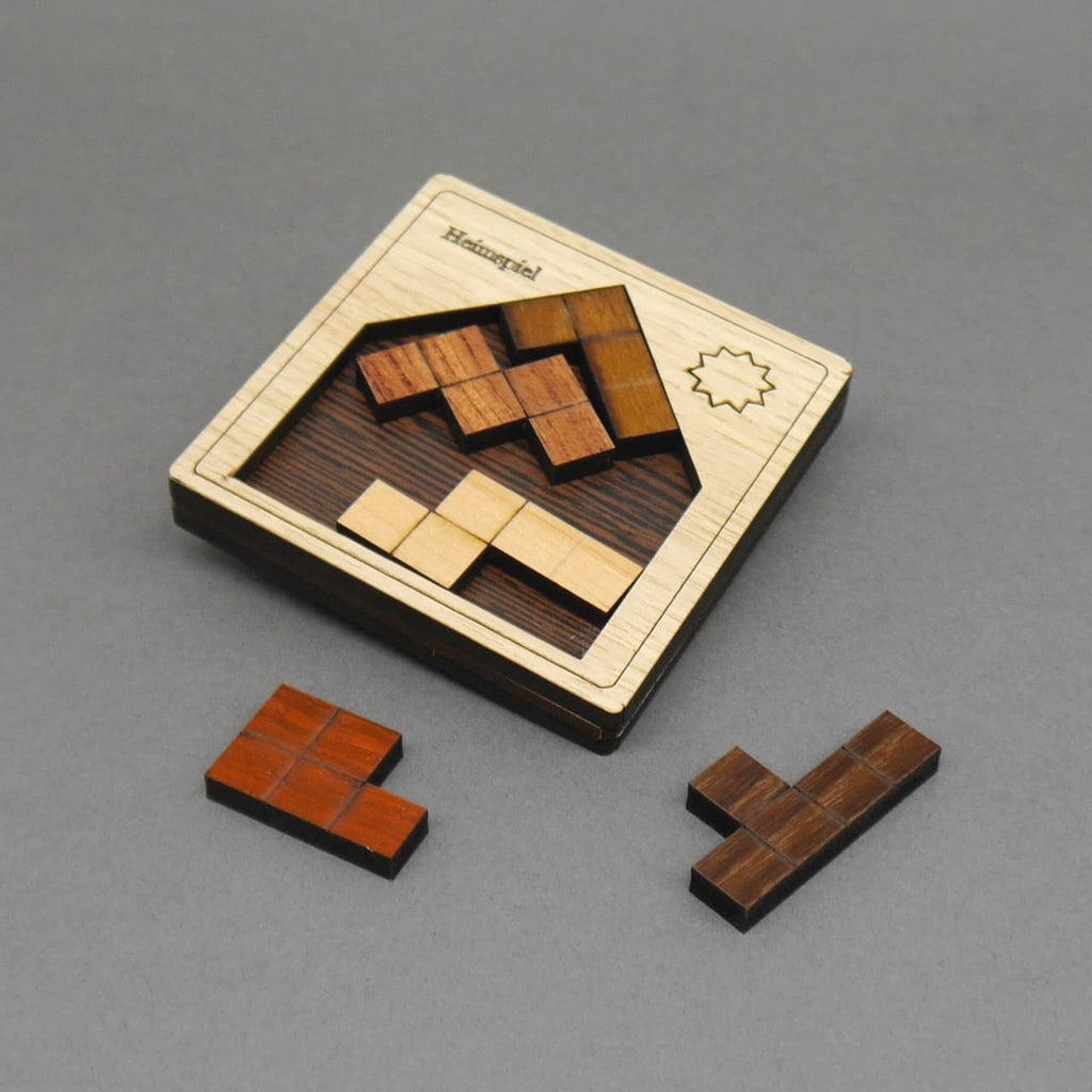 Quadratisches Holzpuzzle „Heimspiel“: heller Hausrahmen mit Sonnengravur; braune und helle Tetrissteine, zwei liegen noch davor.