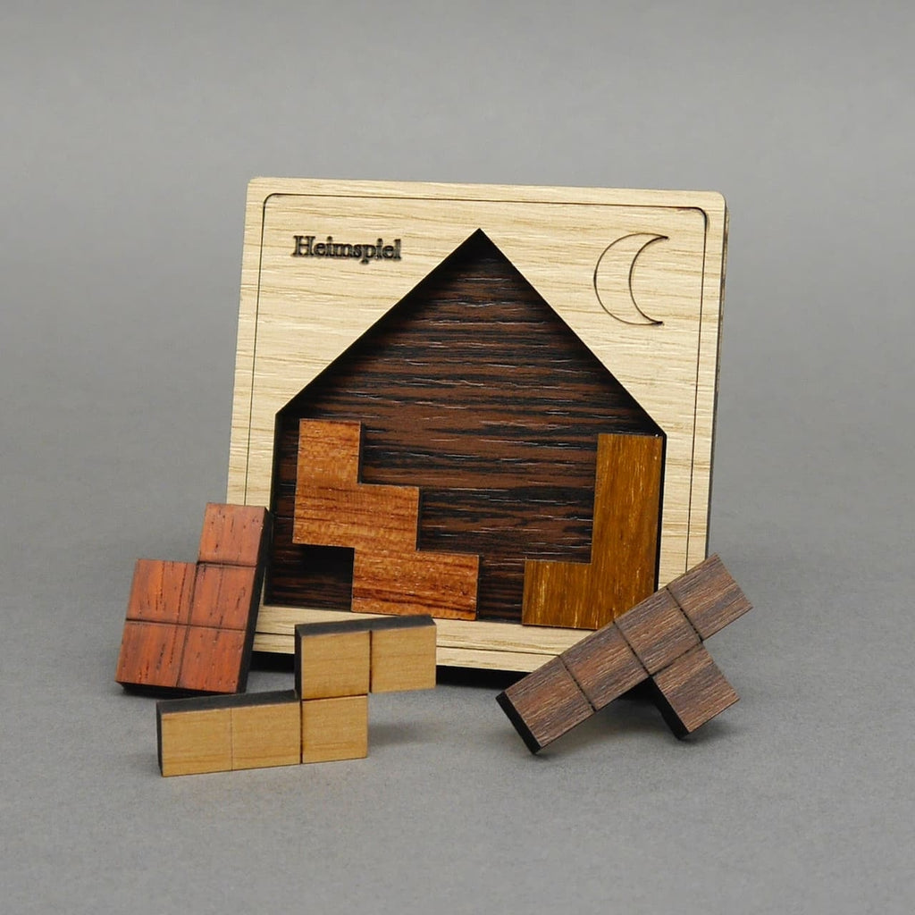 Holzpuzzle „Heimspiel“ in Hausform mit Mondgravur; drei Steine stecken im Rahmen, drei weitere liegen in warmen Holztönen davor.