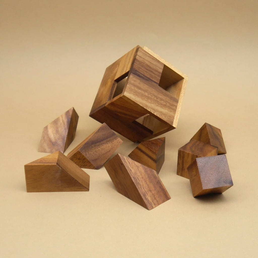 Auseinandergebautes Holzpuzzle „Hexahedroom“ mit hohlem Würfel und verstreut liegenden, teils schräg geschnittenen Holzbausteinen auf beigem Hintergrund.