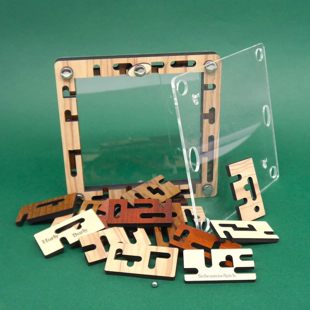 Quadratischer Holzrahmen des Labyrinthspiels „Hurly Burly“ mit abgenommener Acrylabdeckung; zahlreiche verschiedenfarbige Holzplättchen und eine kleine Stahlkugel liegen verstreut auf grünem Untergrund.
