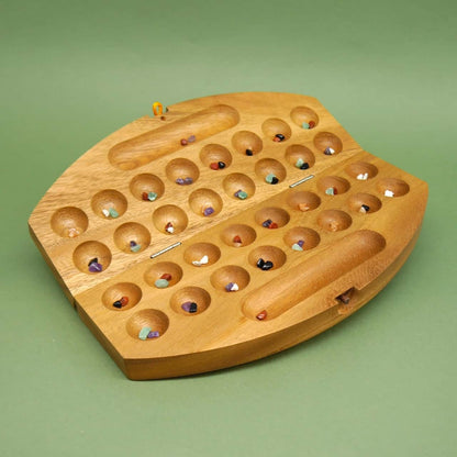 Geöffnetes Mancala-Holzspiel mit farbigen Steinchen in 32 runden Mulden auf grünem Untergrund.