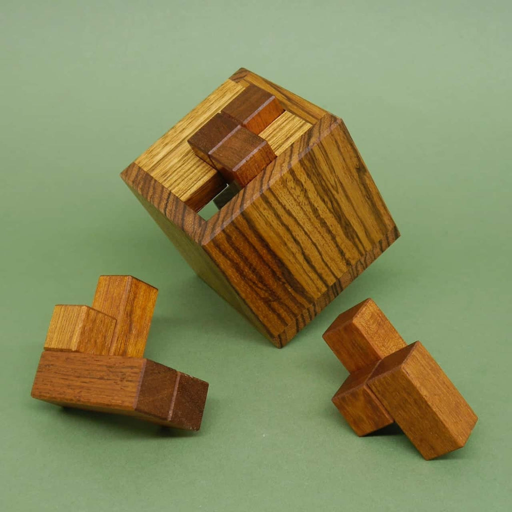 Schräg stehendes Holz-Geduldspiel in Würfelform, drei separate Puzzle-Teile liegen davor auf grünem Untergrund.