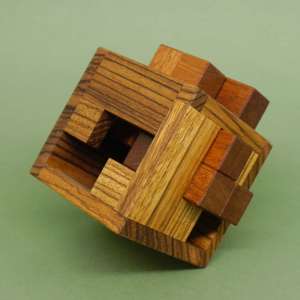Komplexes Knobelspiel: mehrfarbiger Holz-Würfel-Puzzleblock, seitlich geneigt, zeigt freiliegende Steckelemente auf grünem Untergrund.