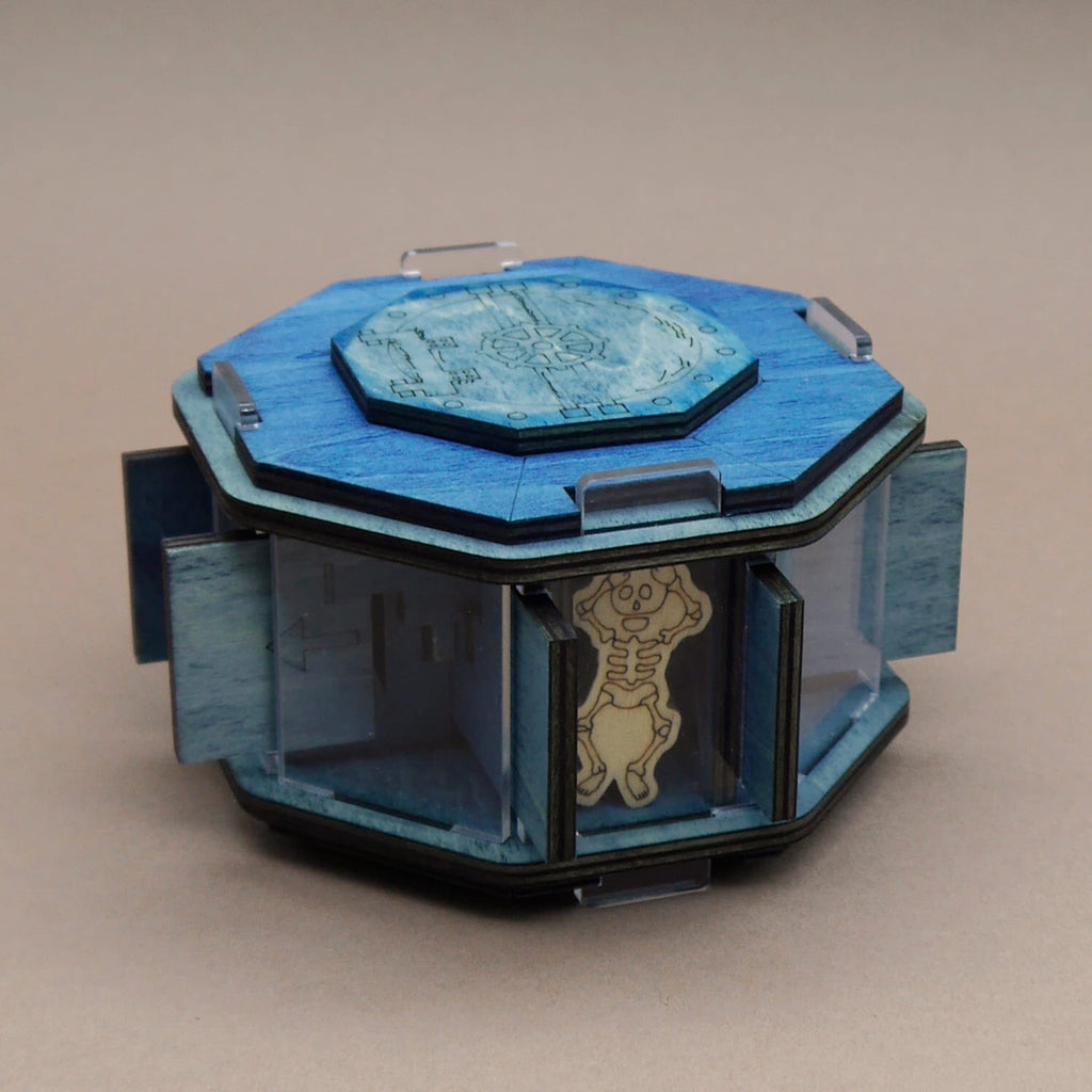 Jailbreak Geheimfach: blaue achteckige Holzbox mit Zahnrad-Deckel und graviertem Skelett hinter Acrylglasfach.