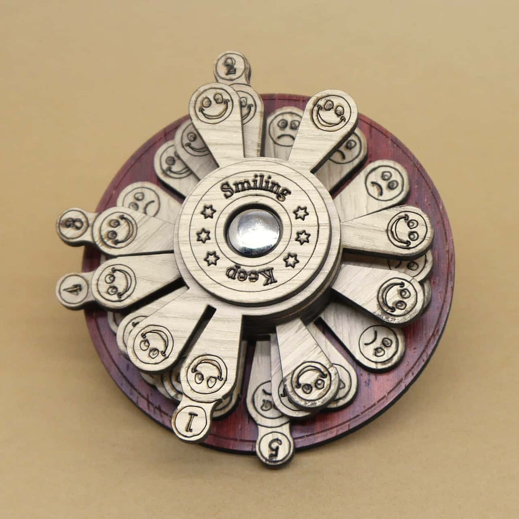 Rundes, mehrschichtiges Holzpuzzle mit mehreren drehbaren Lamellen, auf denen verschiedene Emoji-Gesichter und Zahlen eingraviert sind. In der Mitte ist ein metallener Knopf sowie der Schriftzug „Keep Smiling“ zu sehen.