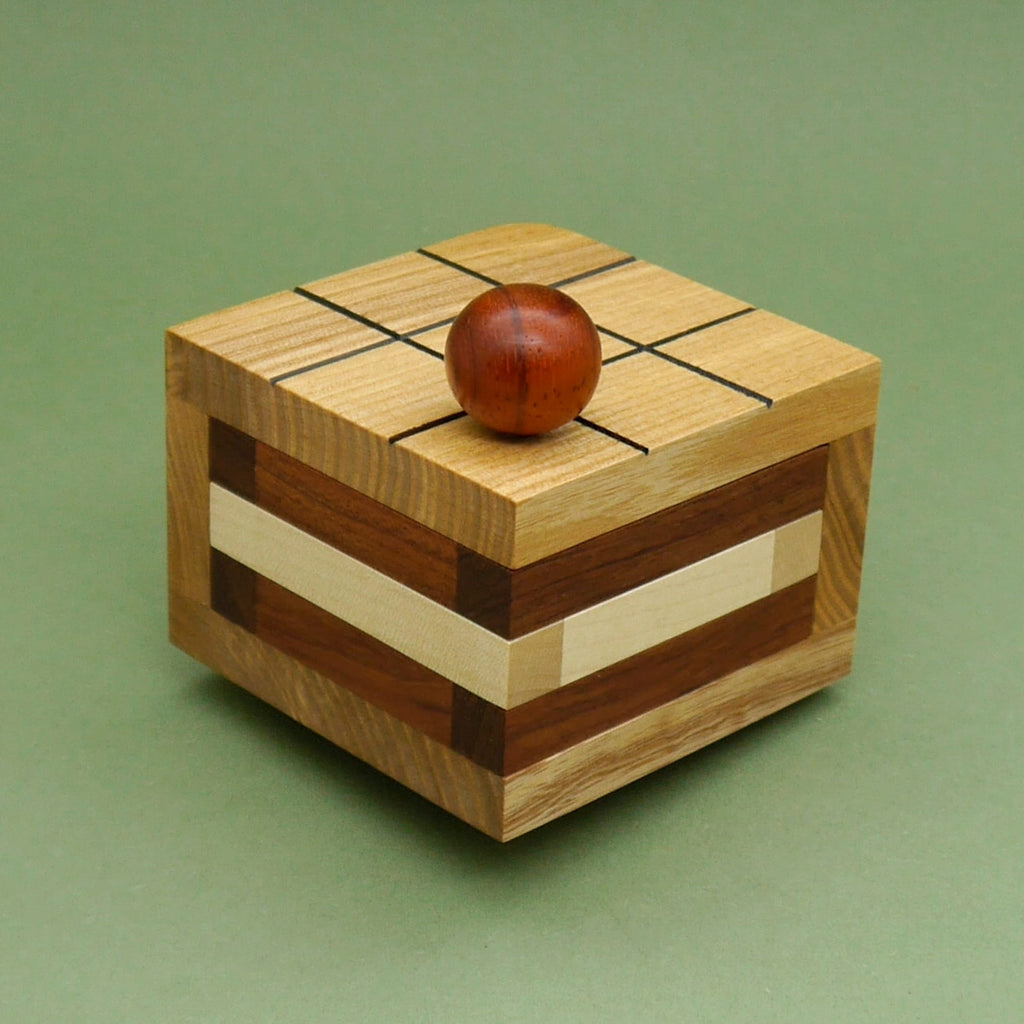 Kuchenstück Holzpuzzle in Würfelform mit geripptem Rasterdeckel, brauner Holzkugel obenauf und gestreiften Seitenflächen.