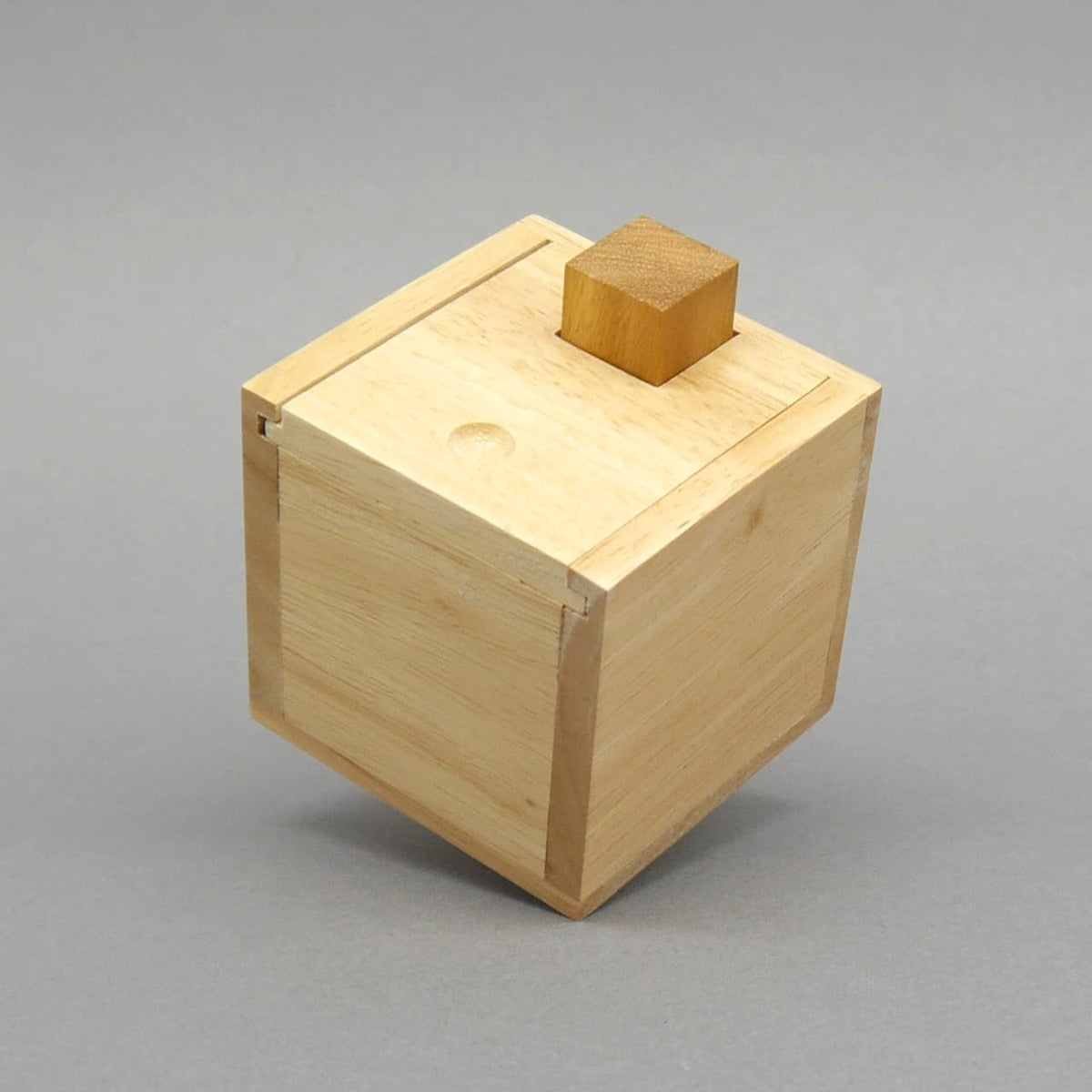 Geschlossene Packpuzzle-Holzbox in Würfelform, Deckel mit aufgesetztem Kubus ragt leicht hervor, freigestellt vor grauem Hintergrund.