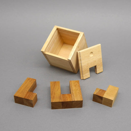 Kleines Logikpuzzle aus Holz: offene Würfelbox, Deckel und drei L-förmige Holzklötze liegen sortiert auf grauer Unterlage.