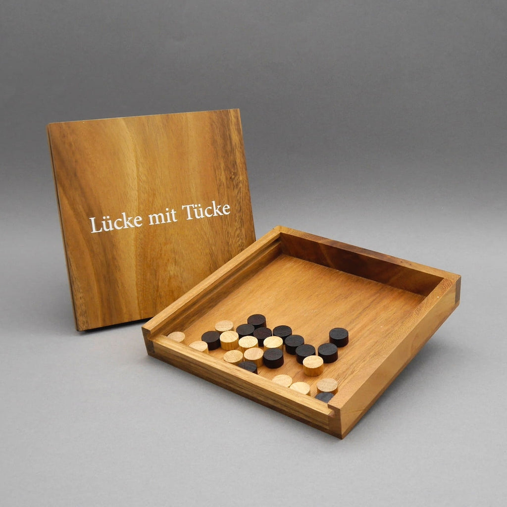 Geöffnetes Reisespiel „Lücke mit Tücke“ aus Holz mit helle- und dunkelbraunen Spielsteinen; Deckel mit Titel lehnt im Hintergrund.