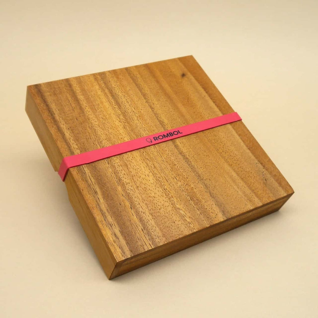 Quadratische Holzbox mit rotem Gummiband als Verpackung für den Spieleklassiker Ludo, schlicht auf beigem Hintergrund.