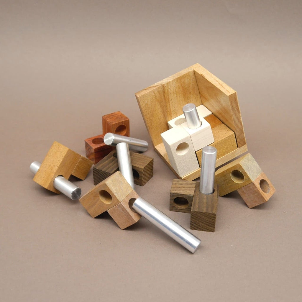 Denkspiel in teilweisem Aufbau – bunte Holzblöcke mit eingelassenen Metallzylindern liegen um ein offenes Holzwürfelgehäuse herum.