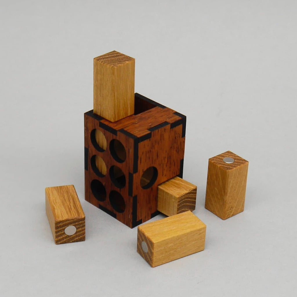 Aufgestelltes Minima Magnetik Packpuzzle: dunkler Holzblock mit runden Öffnungen, aus dem ein heller Holzstein herausragt, umgeben von weiteren magnetischen Holzklötzchen für Denksport und Gehirntraining.