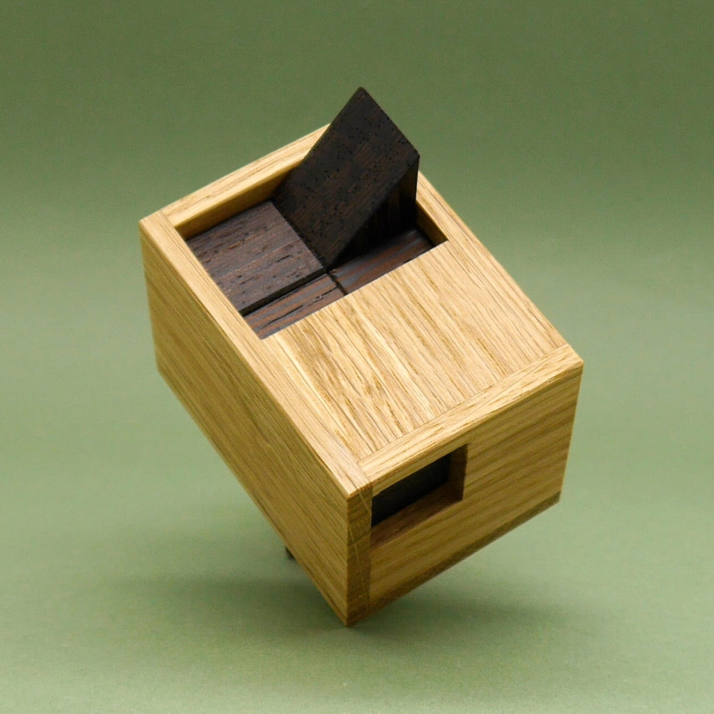 Minima-Ronde-3D-Puzzle auf grünem Grund: aus der hellen Holzbox ragt ein dunkles, keilförmiges Puzzleteil heraus.