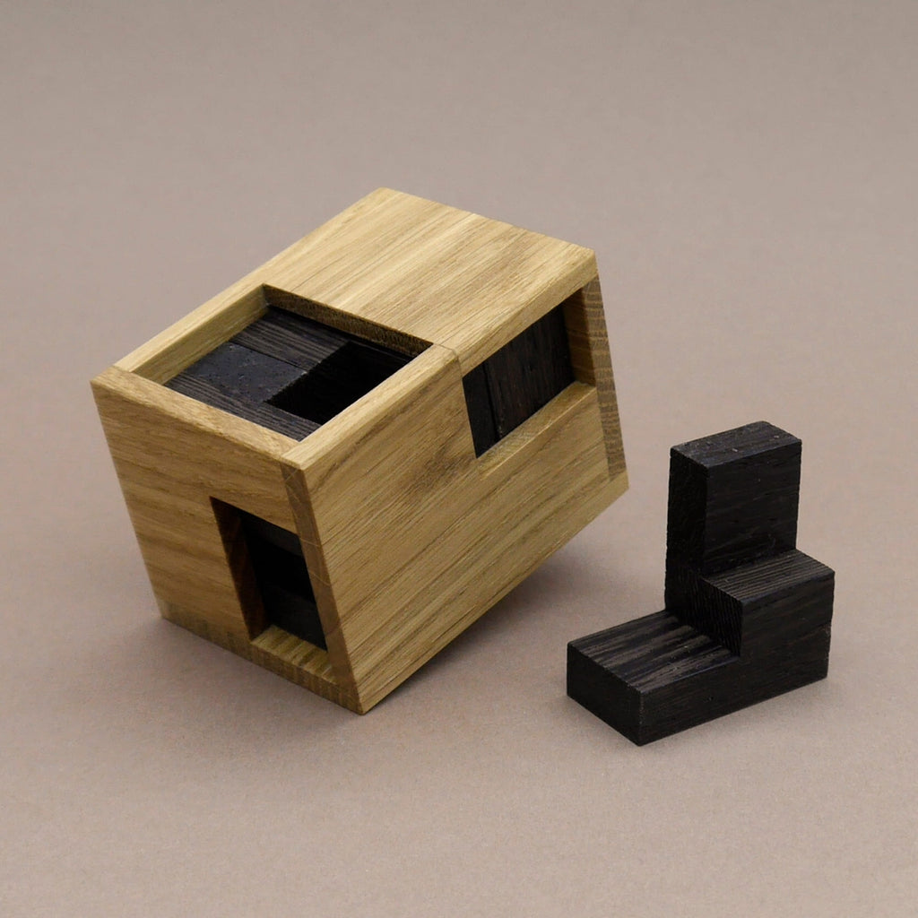 Minima Knobelbox aus hellem Holz mit teilweise eingesetzten dunklen Teilen, ein loses Teil liegt daneben.