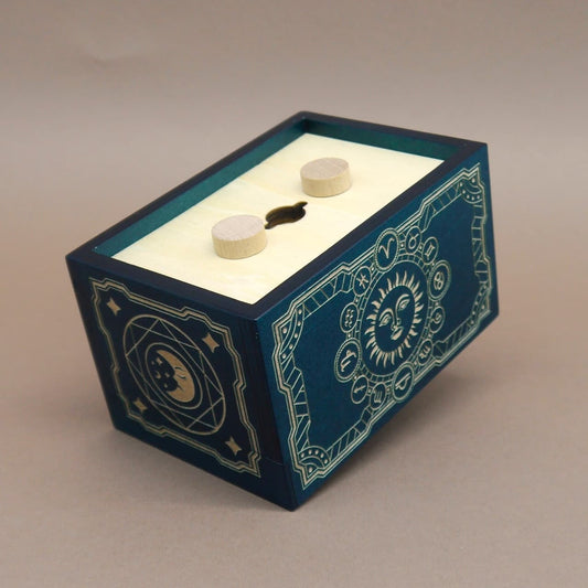 Dunkelblau-türkisfarbene Holz-Puzzlebox mit cremefarbenem Deckel, zwei runden Holzknöpfen und einem schlüssellochförmigen Schlitz; die Seiten tragen goldene astrologische Ornamente mit Sonne-und-Stern-Motiv.