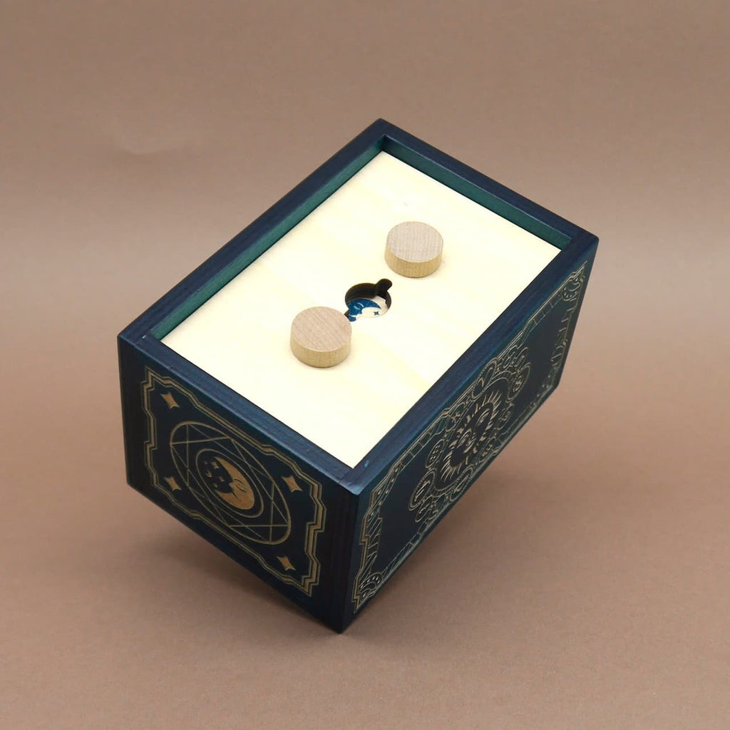 Geschlossene Puzzlebox mit cremefarbenem Deckel und zwei Holzknöpfen; durch den zentralen Schlitz ist ein kleiner, blau glänzender Inhalt als Geheimfach-Überraschung erkennbar.
