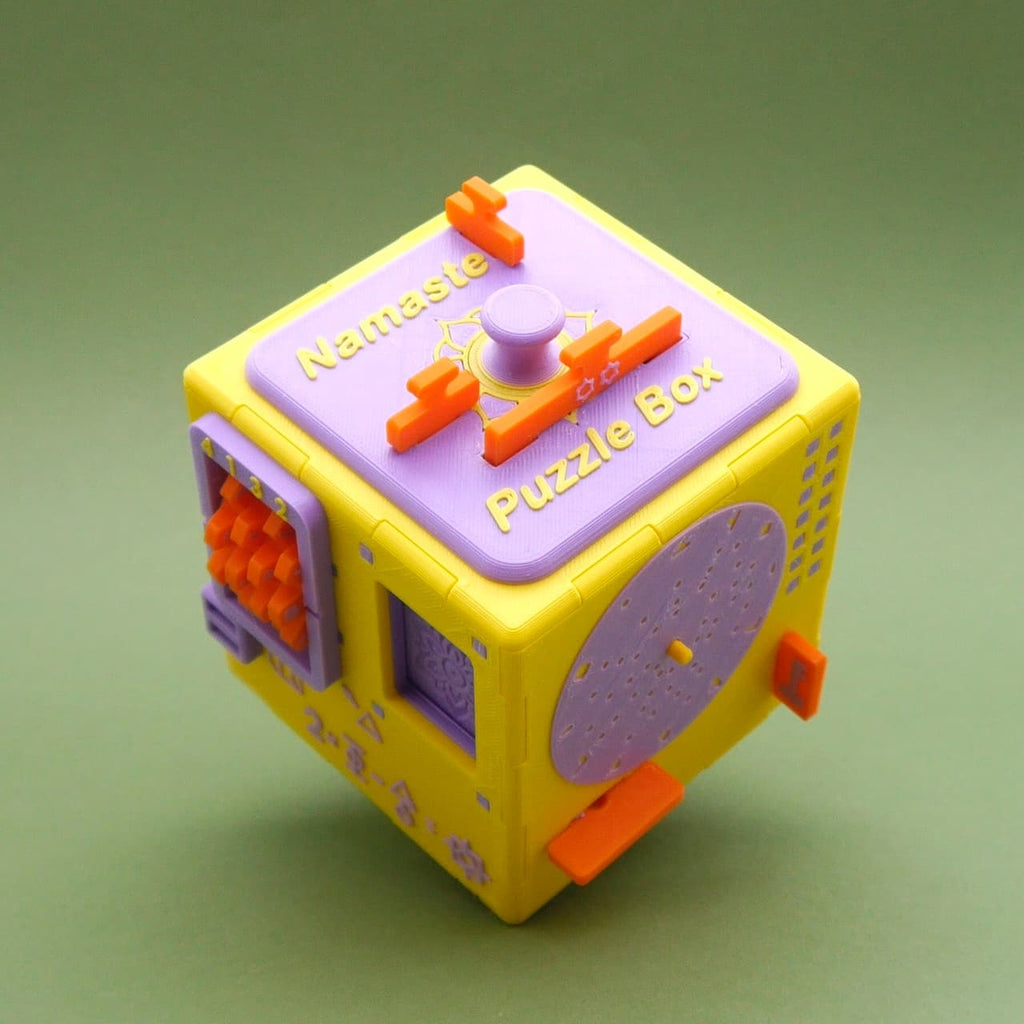 Gelb-lila Namaste Puzzle Box, detailreiche Cluebox mit Schiebern und Drehelementen auf grünem Hintergrund.