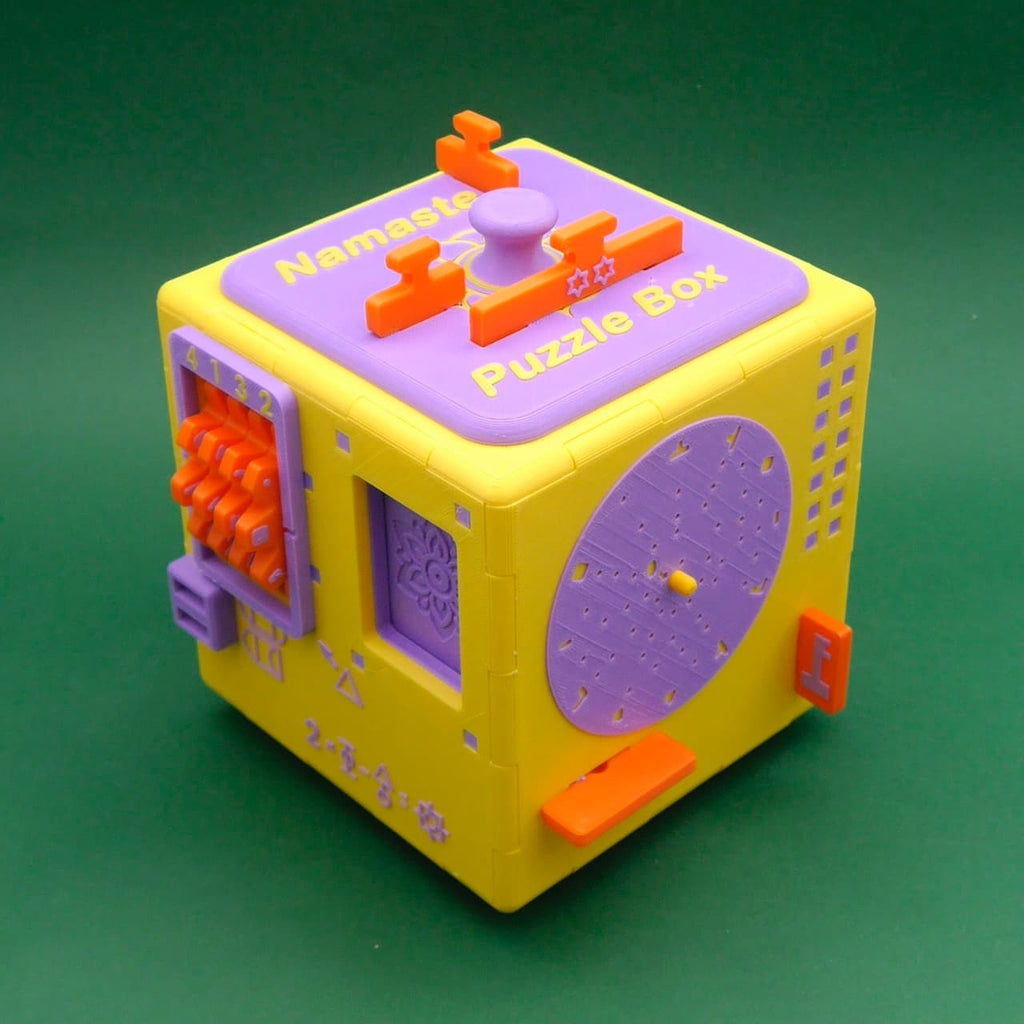Gelb-lila Namaste Puzzle Box als Escape Room Puzzle mit Drehscheibe, Schiebereglern und Codeschloss auf grünem Hintergrund.