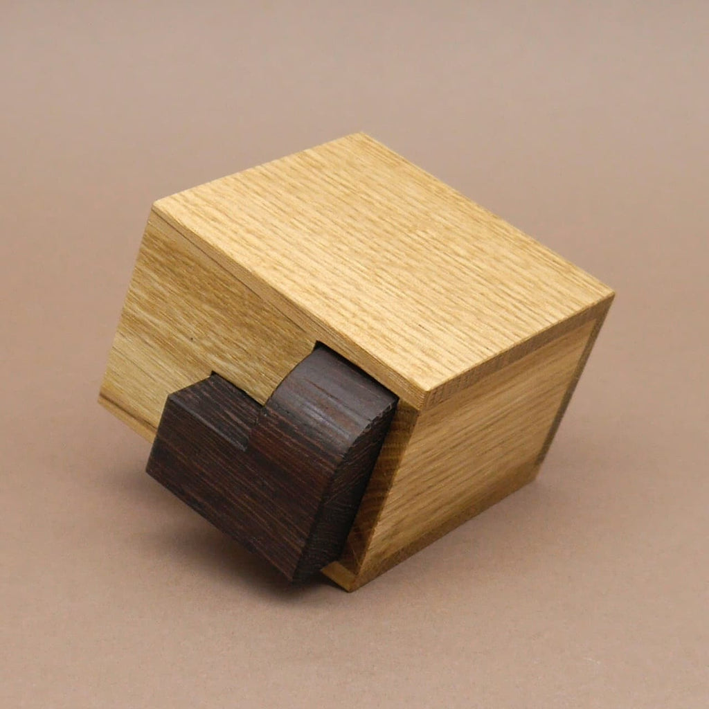 Holz-Denkspiel „Petit Albert“: kleines eckiges Kästchen aus hellem Holz mit dunkelbraunem Puzzleteil, das in die Öffnung geschoben wird.