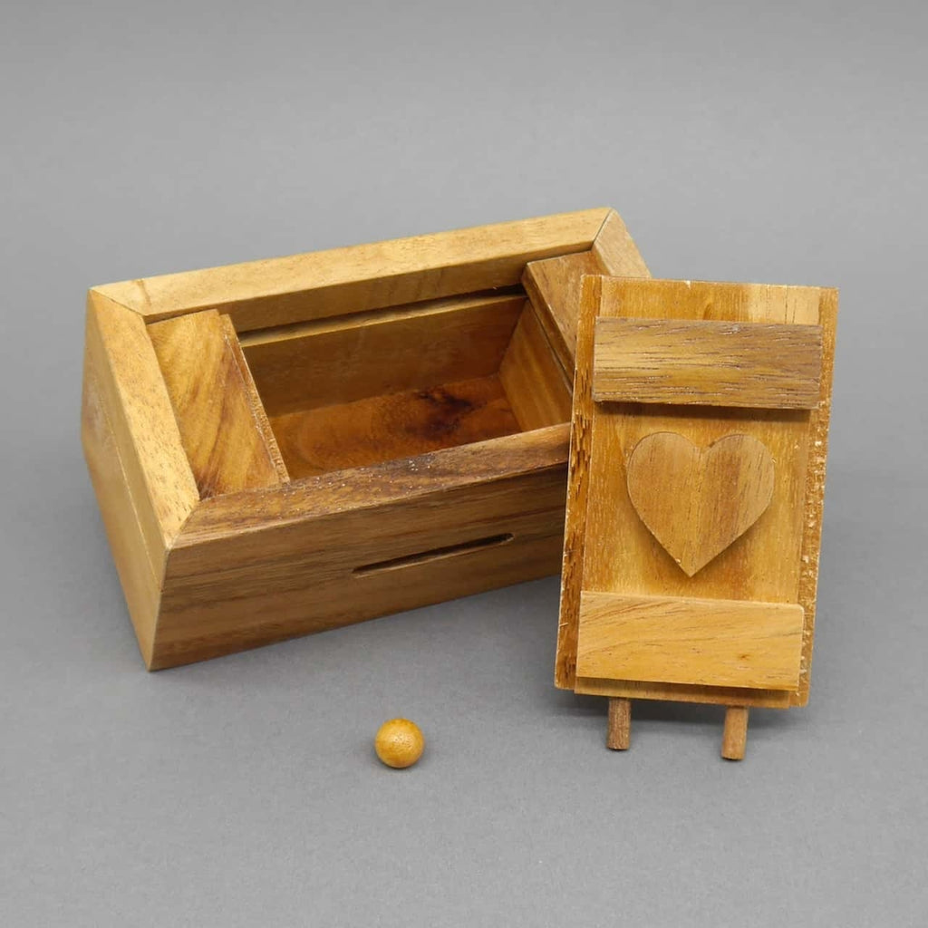 Geöffnete Holzbox eines Geduldspiels, Deckel mit Herzsymbol daneben, kleine Holzkugel davor.