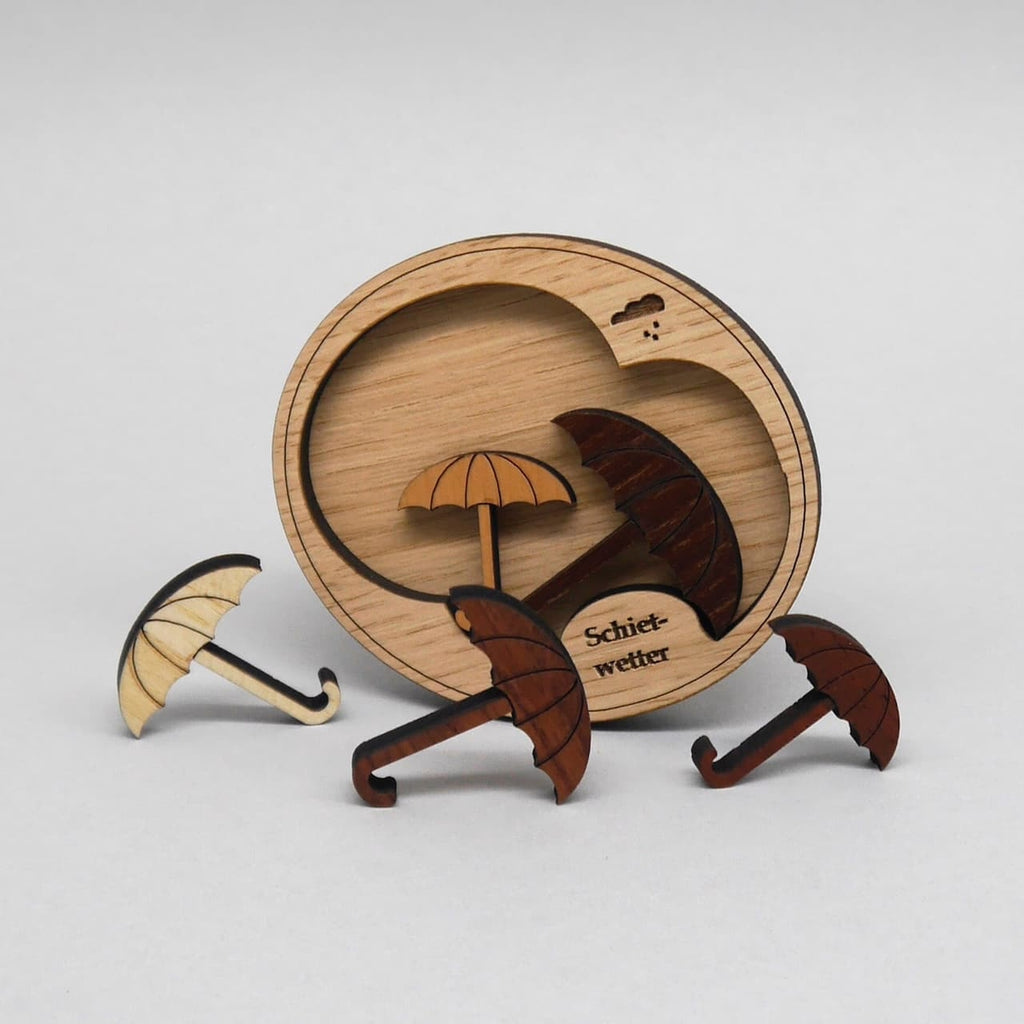 Holz-Denkspiel „Schietwetter“: Runde Wolken-Basis mit herausnehmbaren Schirm-Puzzleteilen aus verschieden dunklen Hölzern, teilweise vor der Basis stehend.