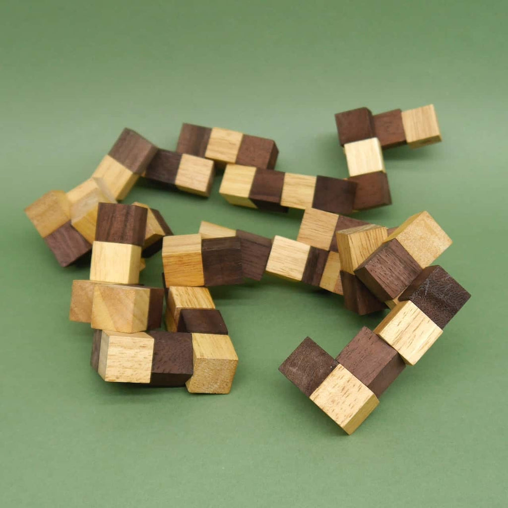 Lang ausgestreckte Holzpuzzle-Schlangenkette aus hell- und dunkelbraunen Würfeln, locker auf einem grünen Boden liegend.