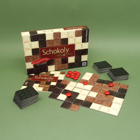 Das Gesellschaftsspiel 'Schokoly' aufgebaut mit Spielbox, Anleitung, Schokoladenplättchen und roten Spielsteinen auf einem grünen Untergrund.