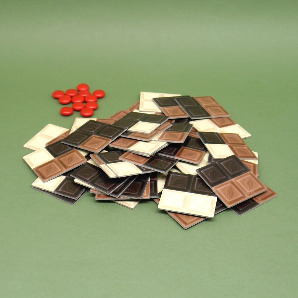 Ein bunter Haufen aus Spielplättchen in Schokoladenoptik auf grünem Hintergrund – das Legespiel zeigt dunkle, helle und weiße Schokotafeln, begleitet von roten Markern.