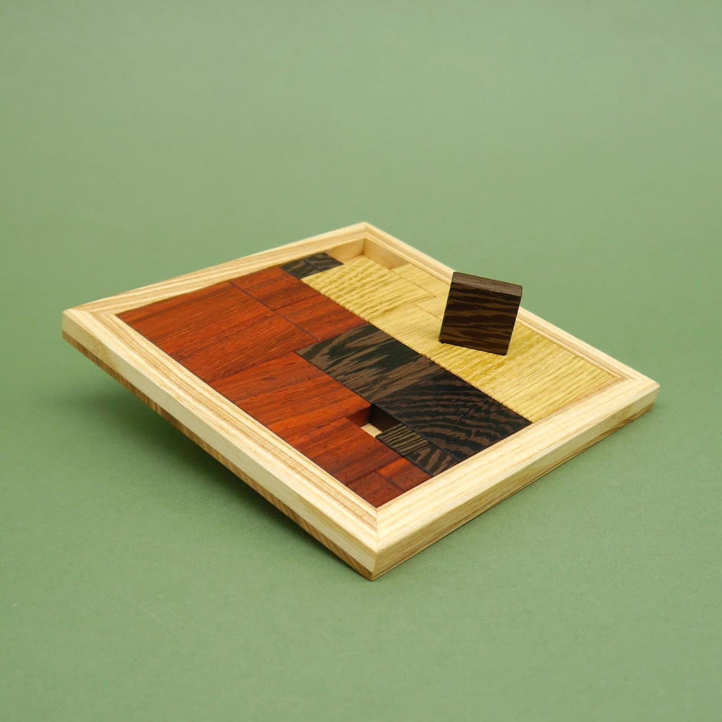 Ein fast vollständig ausgefülltes Holzpuzzle in einem quadratischen Rahmen, bestehend aus verschiedenfarbigen Holzquadraten. Ein einzelnes Teil schwebt leicht über dem Spielfeld.